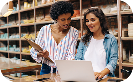 Duas moças em pé próximas a uma mesa olhando para o notebook mostrando algo.