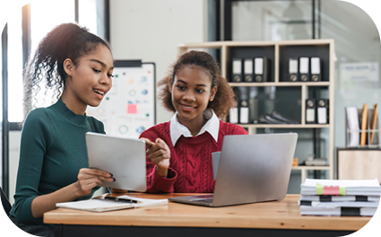 Duas moças sentadas em uma mesa olhando para o notebook com uma expressão de curiosas.
