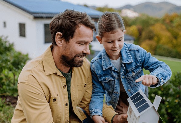 Pai e filha em brincando com uma mini casa com energia solar no teto e ambiente de natureza no fundo.