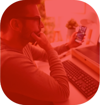 Homem de óculos com uma cara de dúvida, enquanto segura um celular com a mão esquerda e apoia a mão direita no queixo e olha para o seu computador à sua frente.