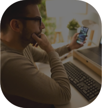 Homem de óculos com uma cara de dúvida, enquanto segura um celular com a mão esquerda e apoia a mão direita no queixo e olha para o seu computador à sua frente.