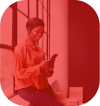 Mulher de camisa laranja e calça jeans apoiada em um parapeito de uma janela sorrindo enquanto olha para o celular.