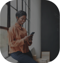 Mulher de camisa laranja e calça jeans apoiada em um parapeito de uma janela sorrindo enquanto olha para o celular.