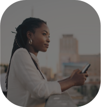 Mulher olhando a paisagem enquanto segura o celular em uma varanda, com alguns prédios desfocados ao fundo.