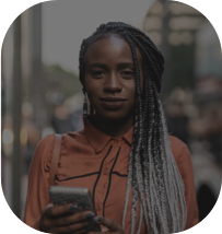 Mulher com uma roupa laranja segurando um celular e com um sorriso de canto e ao fundo prédios.