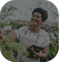 Uma mulher mais velha mexendo nas plantas com um macacão e segurando um tablet na outra em um ambiente de plantação.