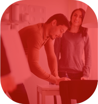 Homem assinando um papel apoiado na mesa e uma mulher parada sorrindo enquanto olha para outro lugar.