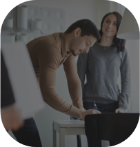 Homem assinando um papel apoiado na mesa e uma mulher parada sorrindo enquanto olha para outro lugar.
