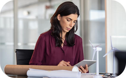 Moça em um escritório com uma roupa de cor vinho mexendo em seu tablet com algumas papeladas em cima da mesa.