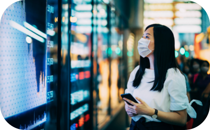 Moça com roupa branca e máscara parada em frente a um telão com diversos números e gráficos e segurando um celular.