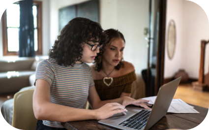 Duas moças sentadas em uma mesa olhando para o notebook com uma expressão de curiosas.